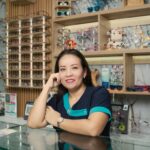 Smiling female optician in eyewear store standing behind counter surrounded by glasses displays.